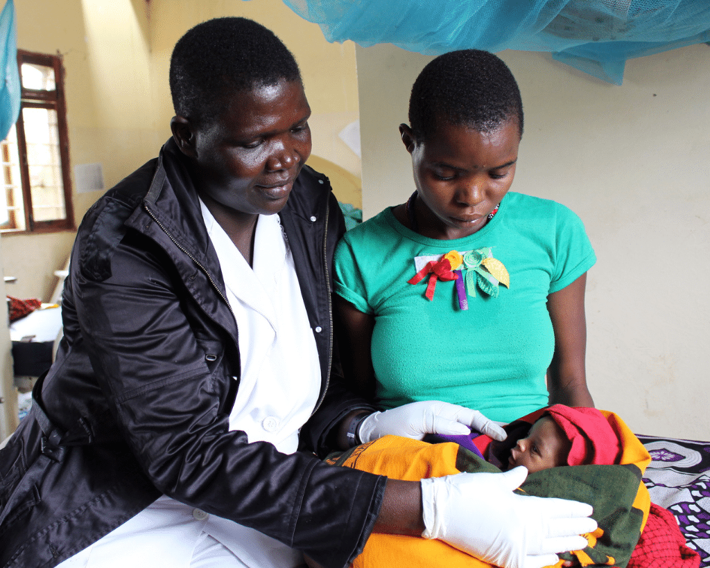 a health worker wearing gloves holds a baby with its mother just behind