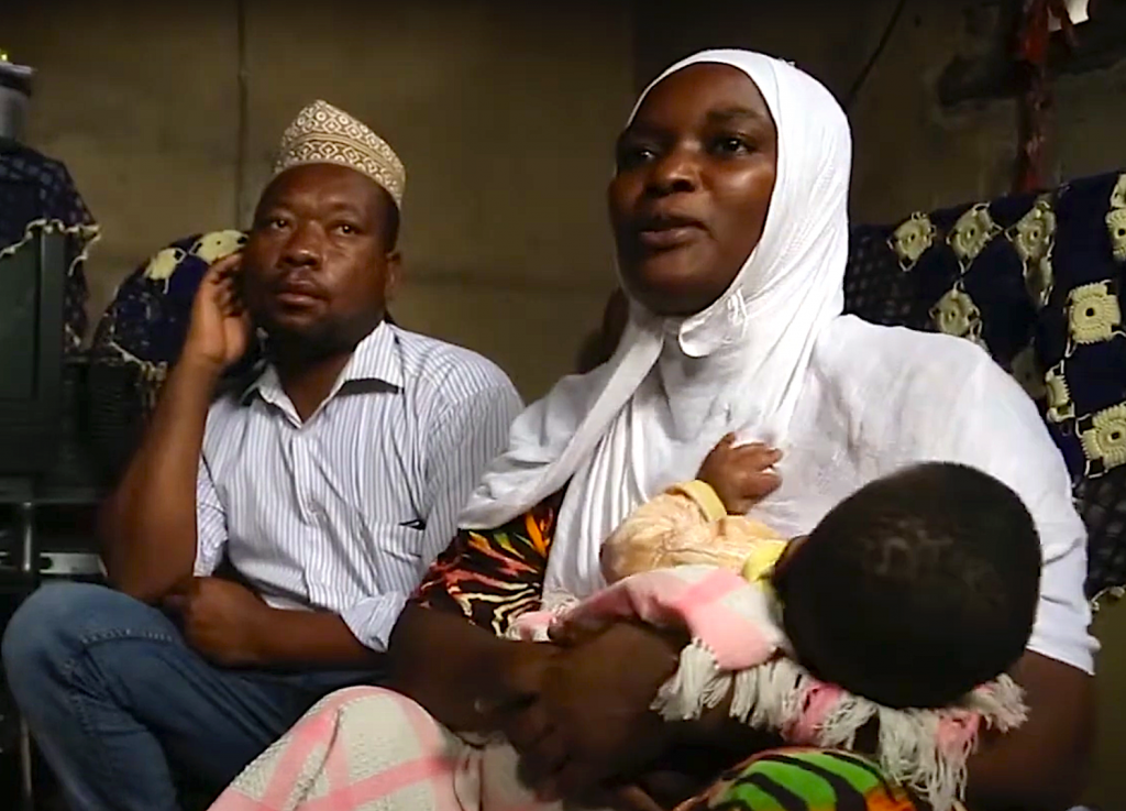 A man and woman sit next to each other. The woman holds a baby in her arms. She wears a white headdress