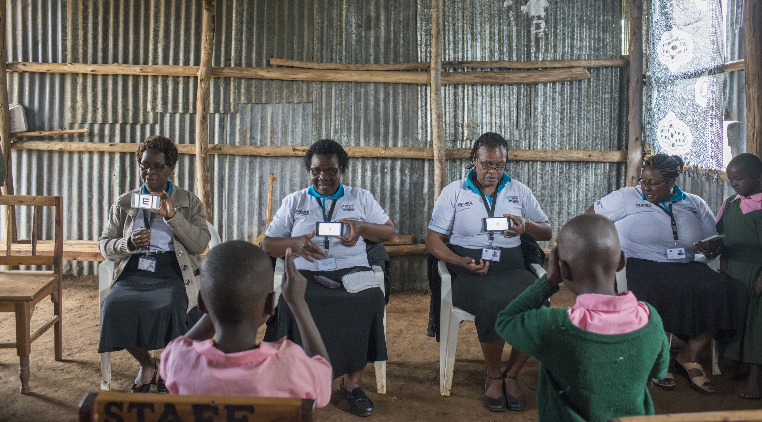 women sit in a row using mobile phones to assess children's sight. The children are sat opposite them