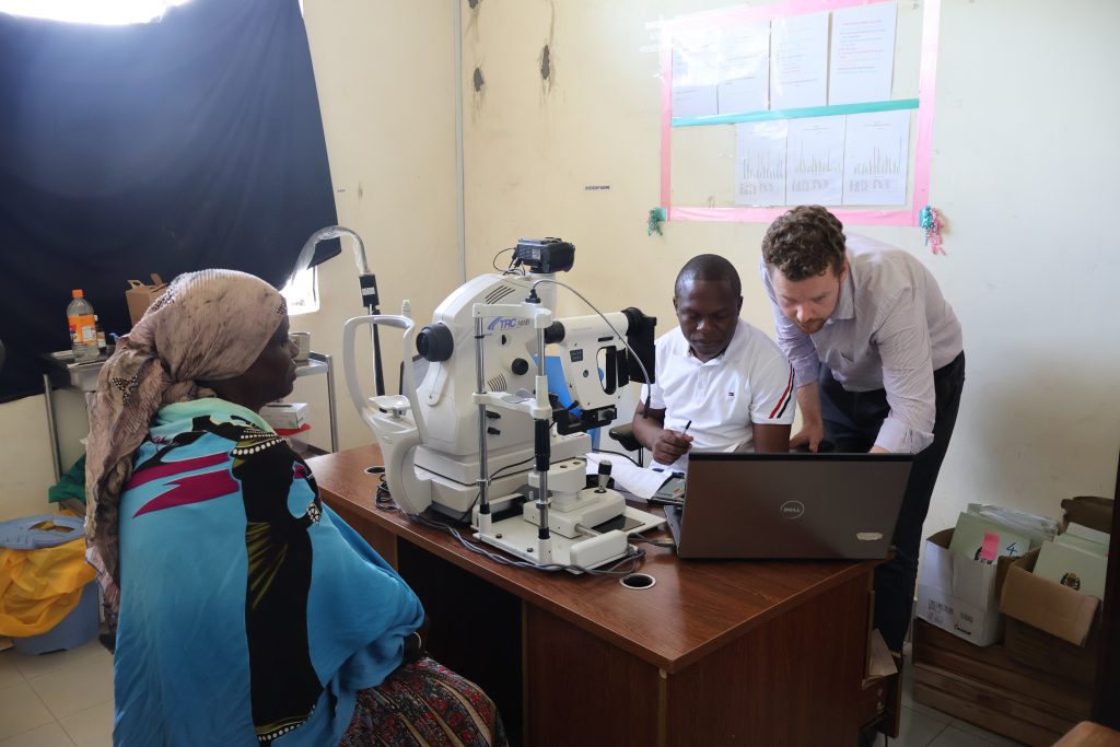 two men work at a laptop while a woman sits behind ophthalmic equipment