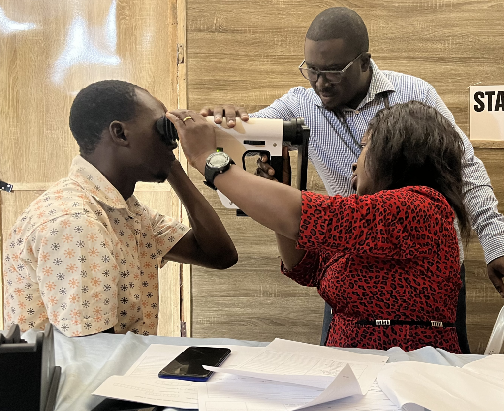a man and a woman hold a handheld fundus camera to another man's eye