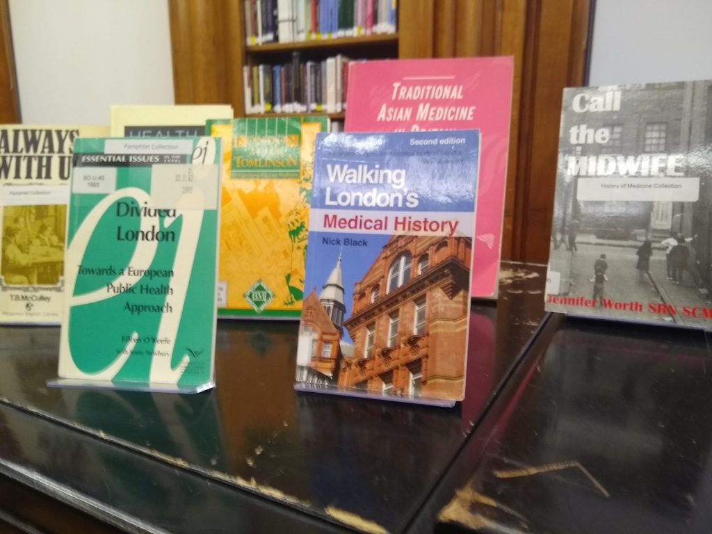 A photograph of half of the display in the Library, consisting of seven brightly coloured books with red, blue, grey, green, yellow, and beige covers. One cover has a photograph of UCL's Cruciform Building, a red-brick Gothic former hospital.
