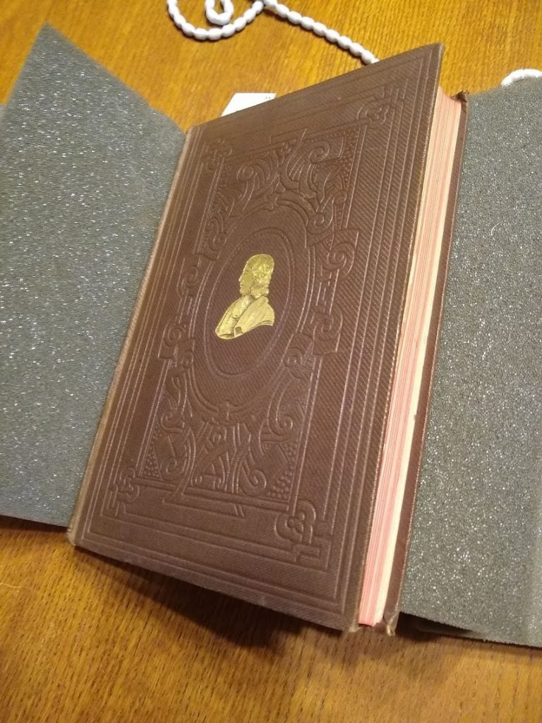 The cover of 'Two monographs on malaria and the parasites of malarial fevers,' in brown, with an embossed pattern of knots, surrounding a gold portrait of the author in profile.