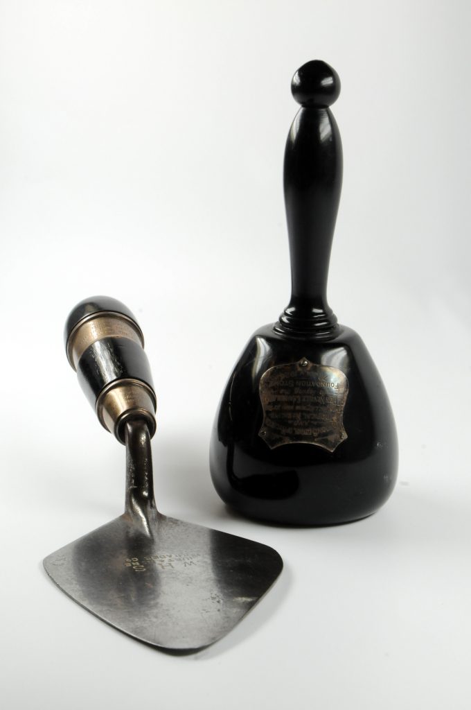 Black mallet with small silver plaque on the front. Beside it is a silver trowel with a black and gold handle. 