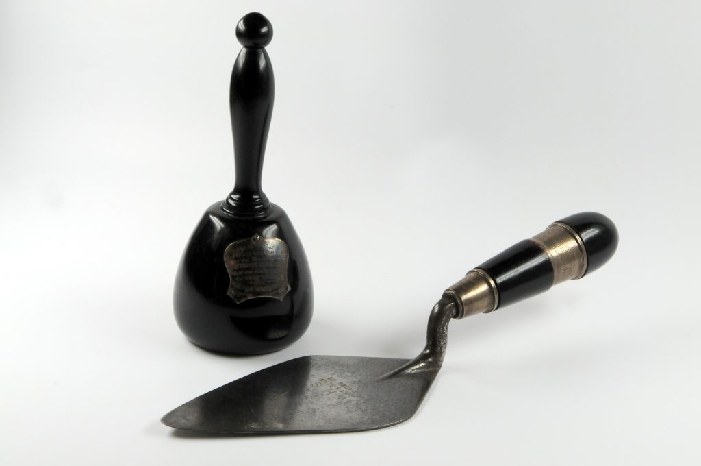 Black mallet with small silver plaque on the front. In front is a silver trowel with a black and gold handle. 