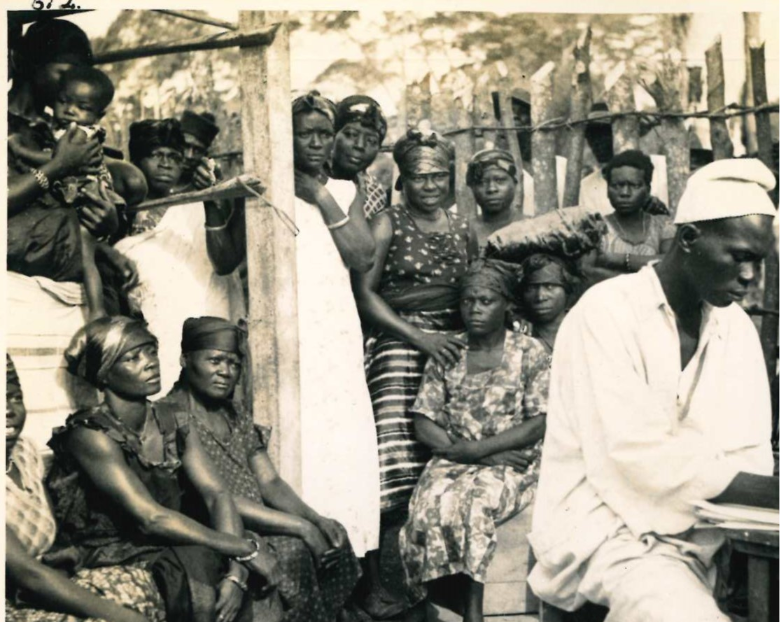 Black and white image of women from Cameroon waiting around a man who is writing in a notebook.